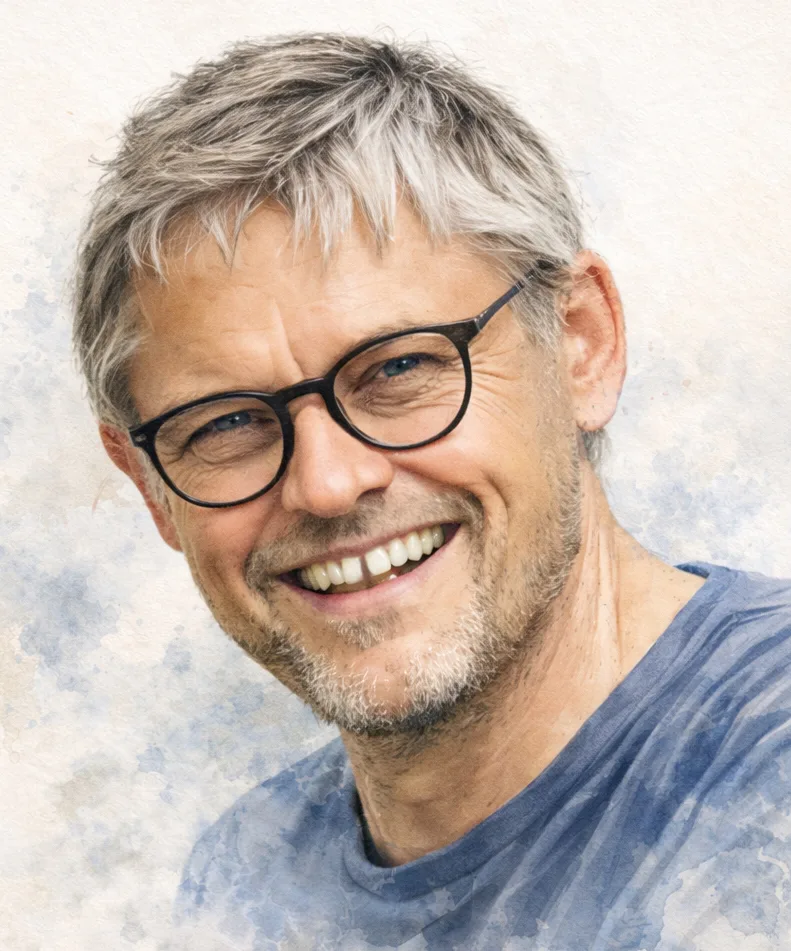 Glimlachende man met grijs haar, bril en stoppelige baard, gekleed in een blauw shirt tegen een lichte achtergrond.
